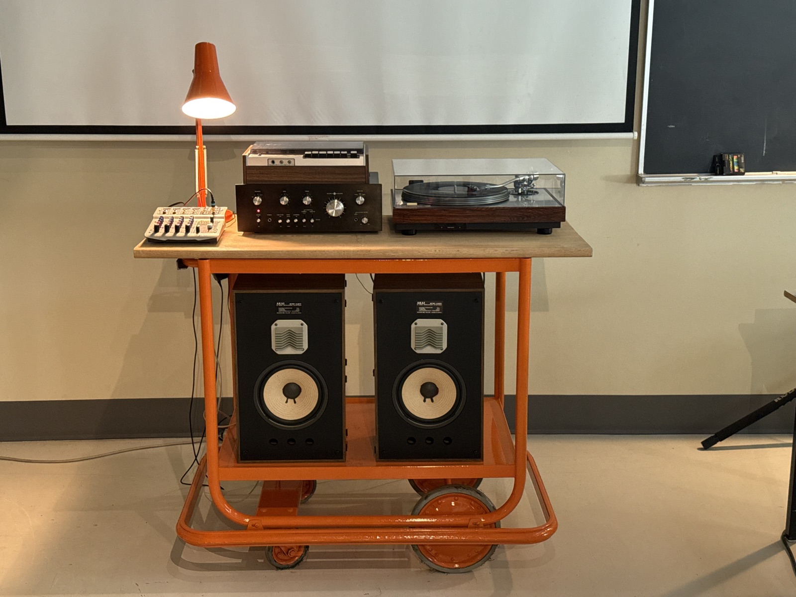 An orange rolling cart in a classroom holding a turntable, amplifier, reel-to-reel deck, mixer and desk lamp, with two Akai speakers on the shelf below.